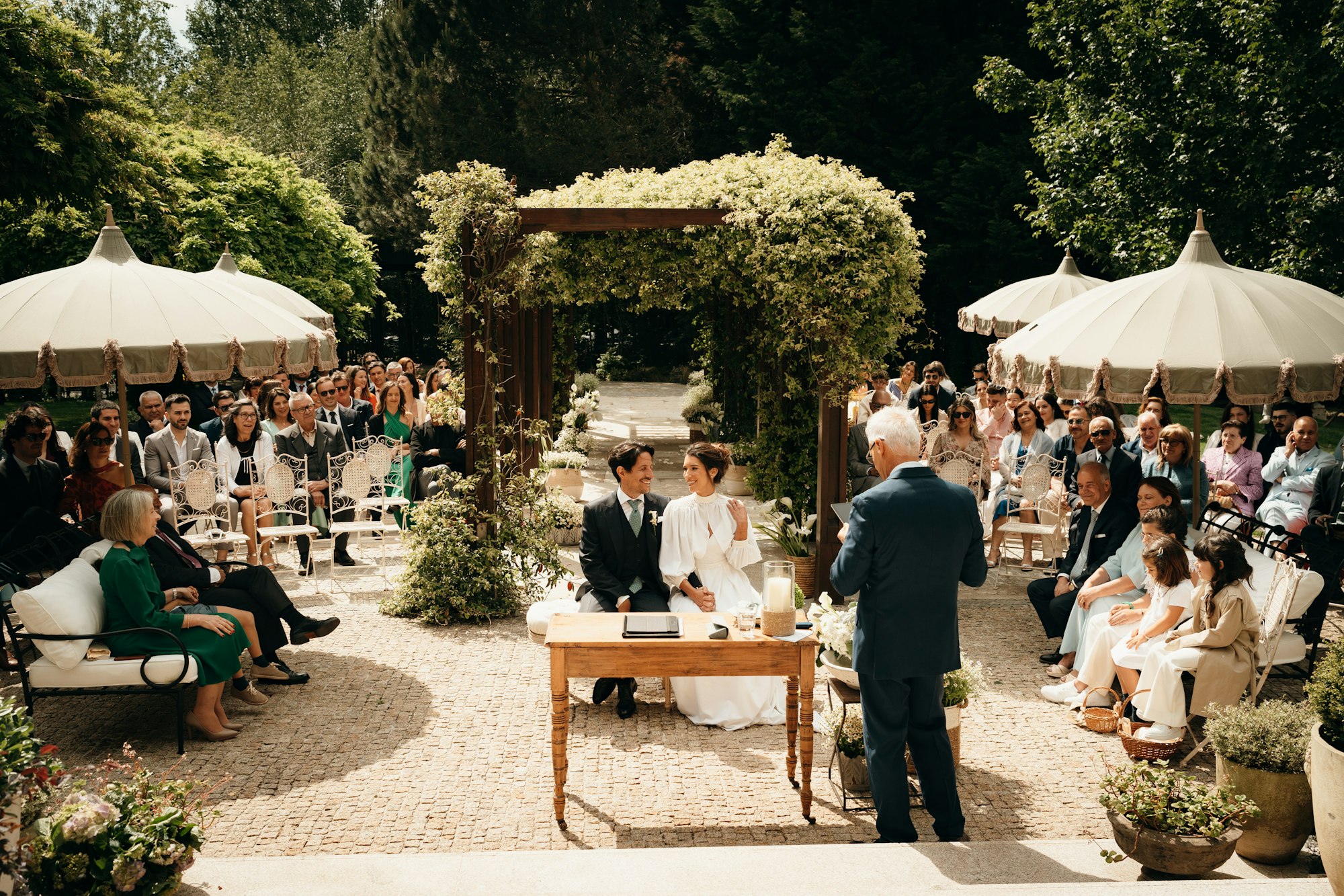 A celebrant standing at the signing table facing the seated couple, guests on both sides of a garden courtyard