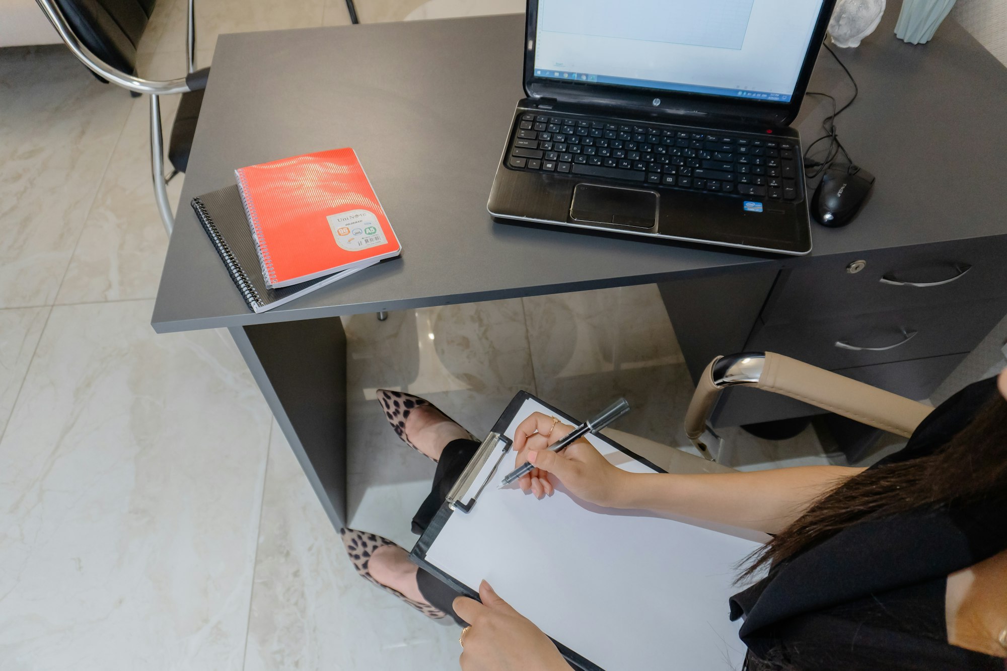 A planner writing on a clipboard at a desk beside an open laptop and paperwork