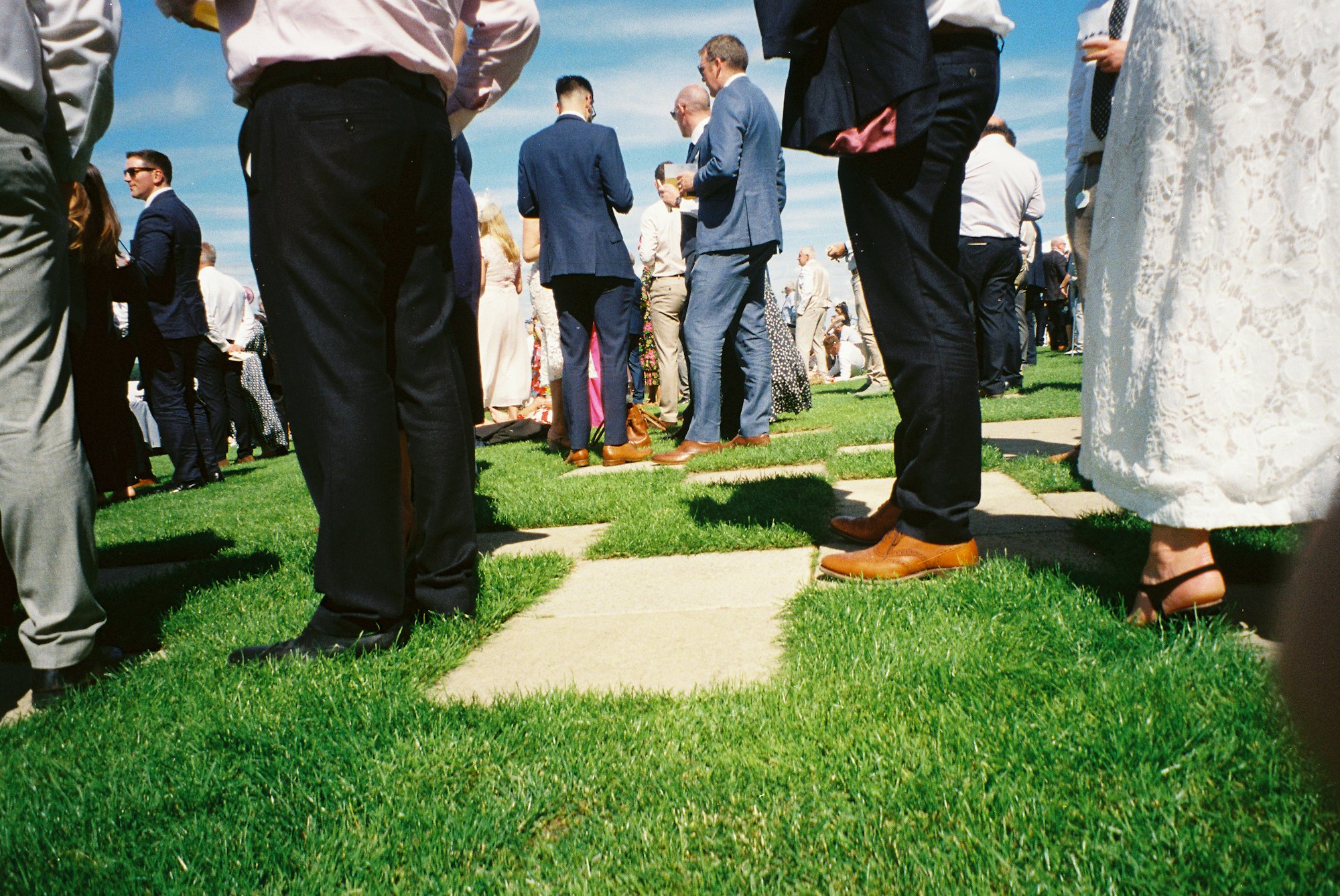 Wedding guests in suits and dresses standing about on a lawn in the sun with drinks in hand, waiting