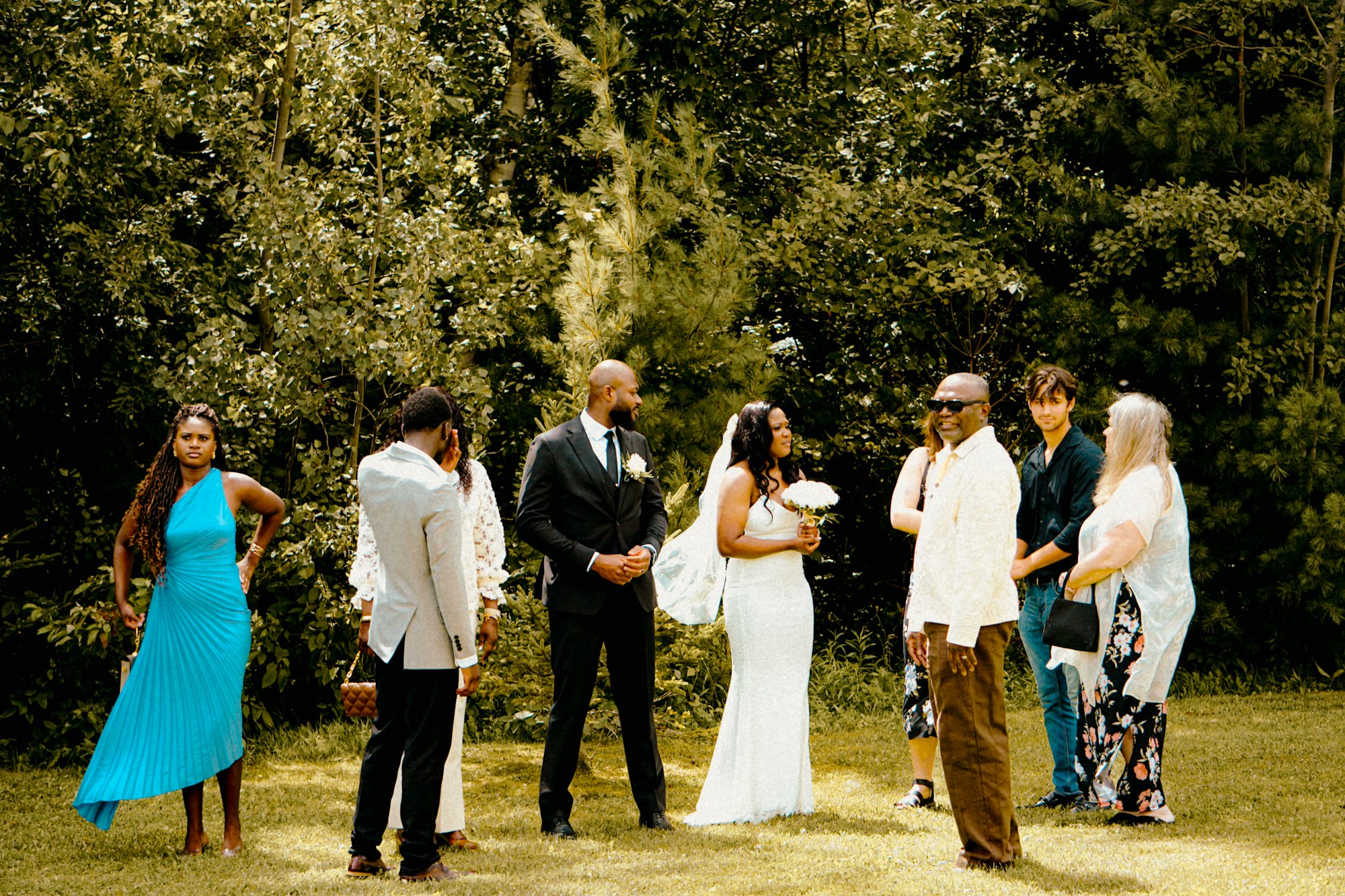 A wedding party standing in a loose line under trees while a group photograph is taken