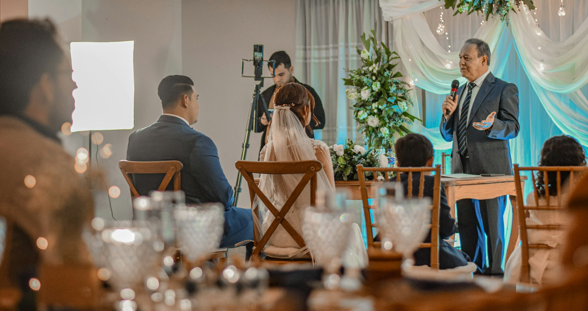 A guest speaking into a microphone at a wedding reception while the couple listen from their table