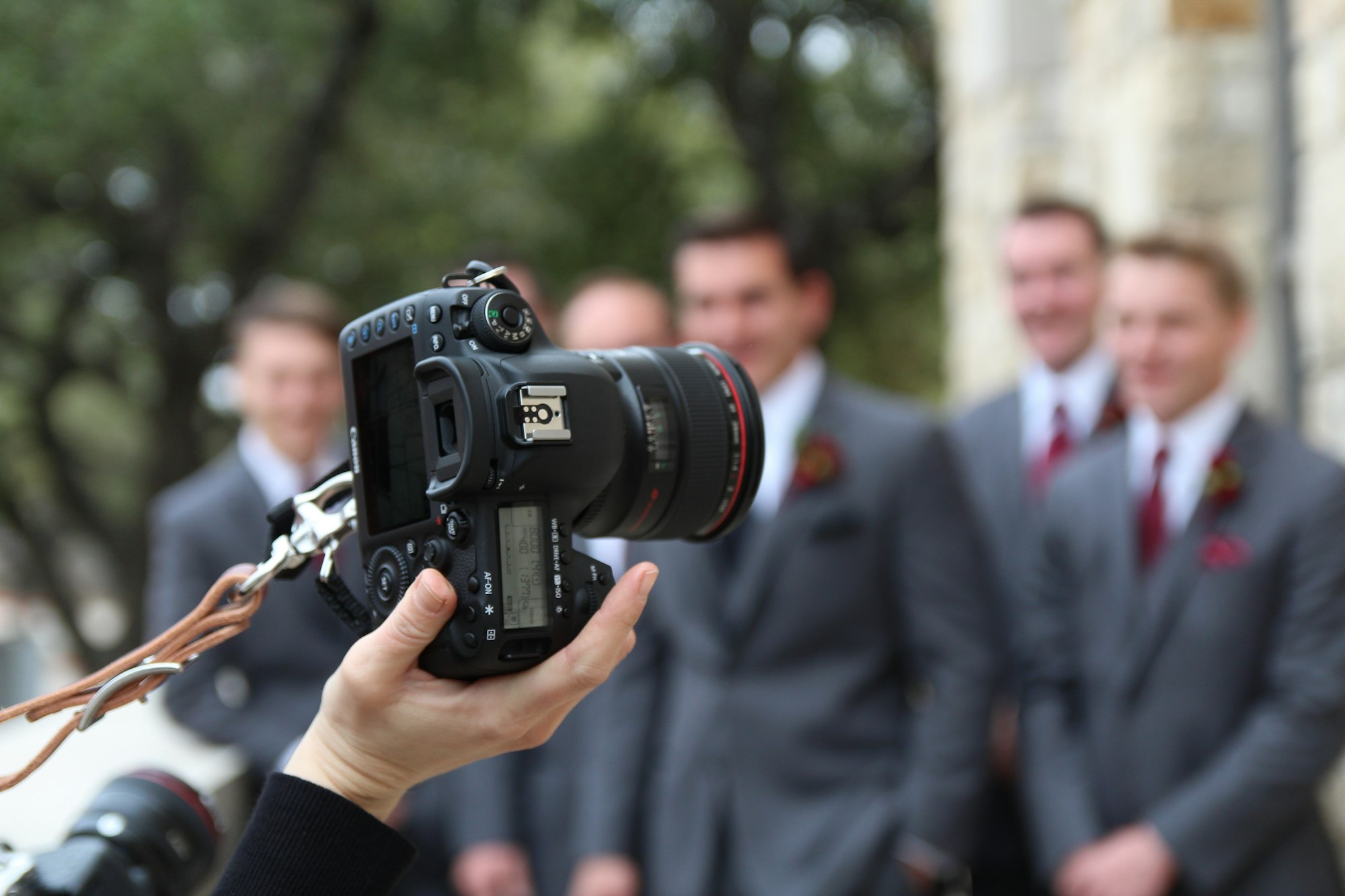 A photographer's camera framing a blurred group of wedding guests in grey suits outside a stone building