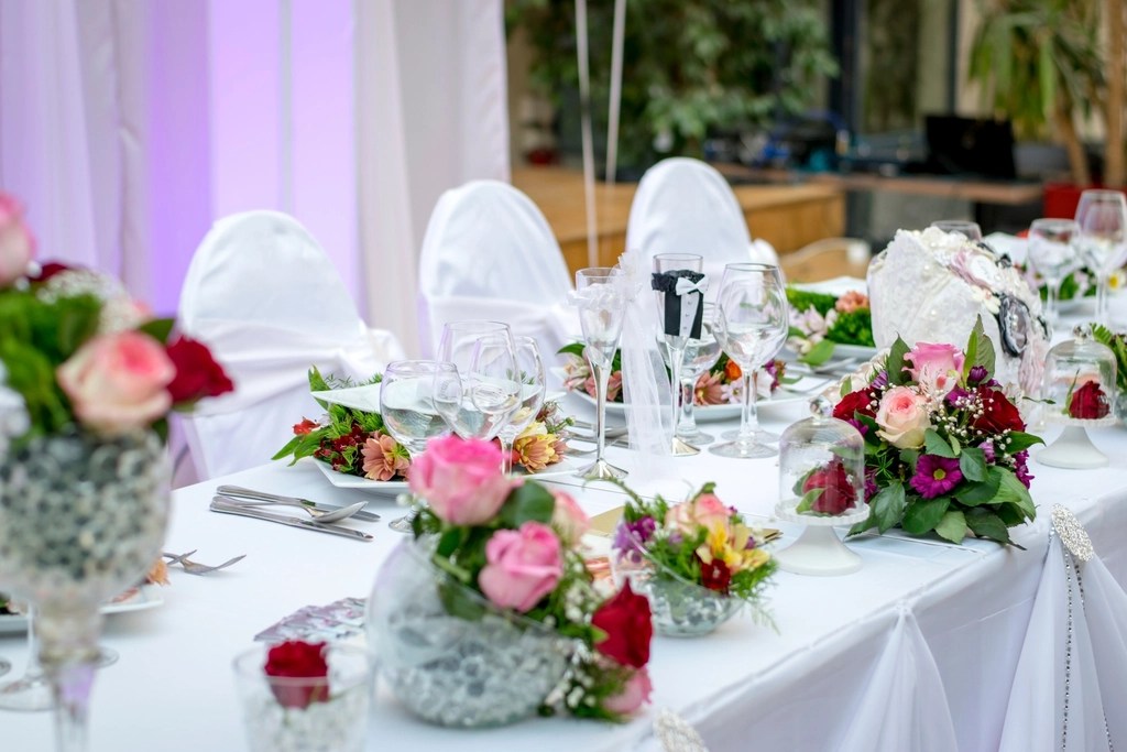 A wedding reception table laid with white linen, rose arrangements and glassware before guests are seated