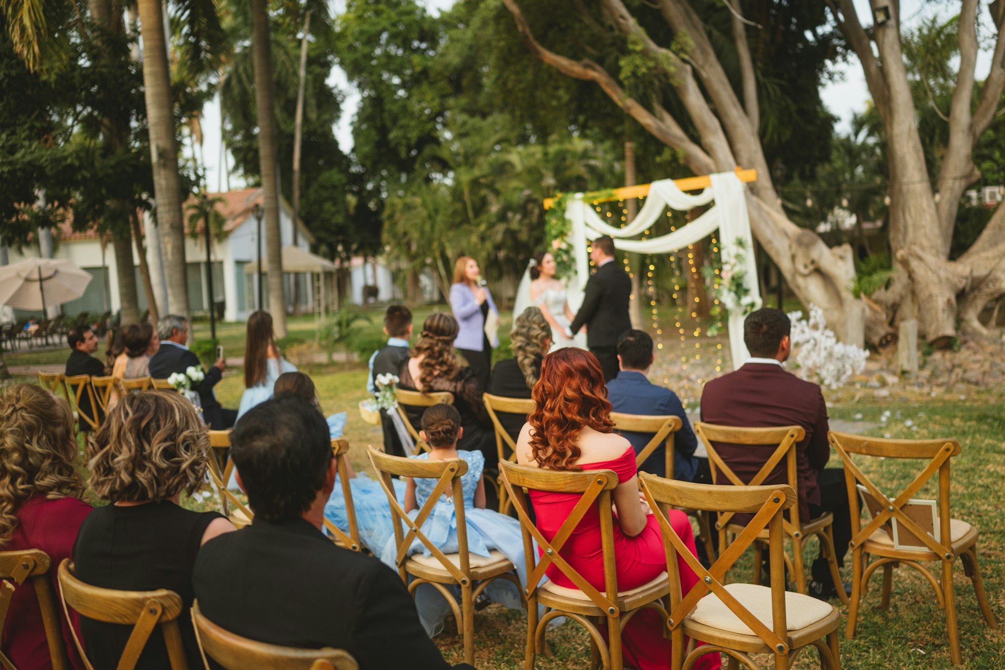 Guests seated on cross-back chairs at an outdoor garden ceremony, the couple at a draped arbour in flat afternoon light