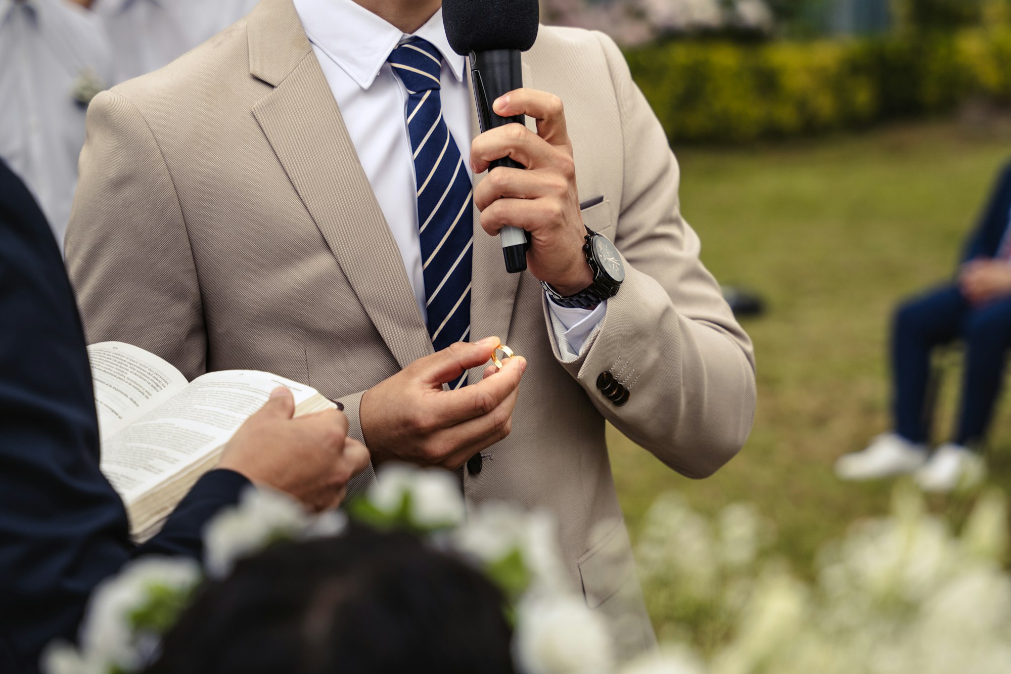 A person in a dark suit holding a microphone and speaking to a room at a reception