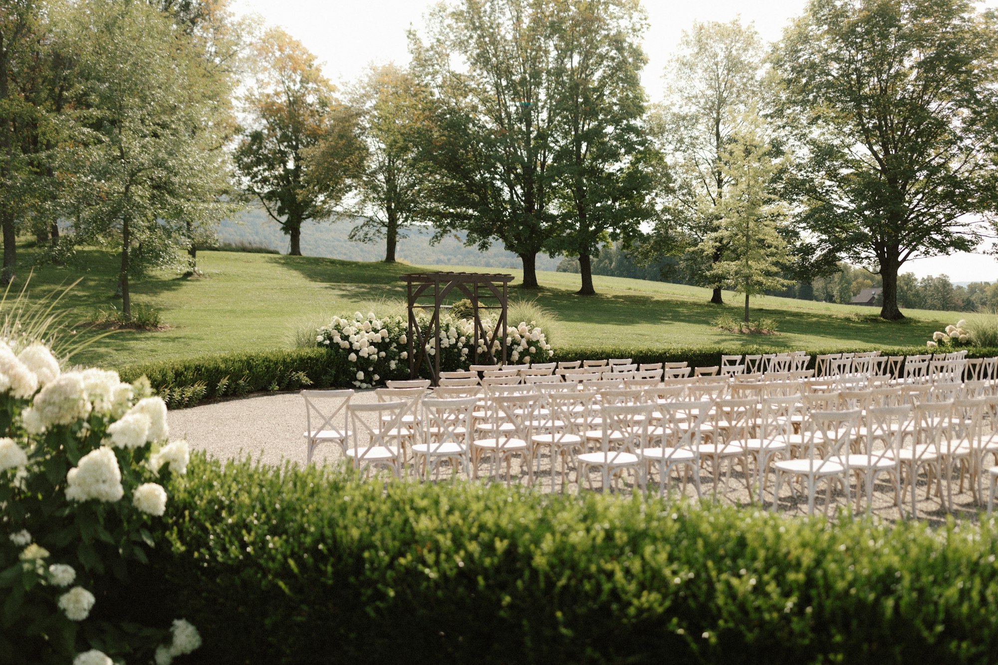 Rows of empty cross-back chairs and a timber arbour set out on a lawn, hedged and flowering, before a single ceremony guest has arrived