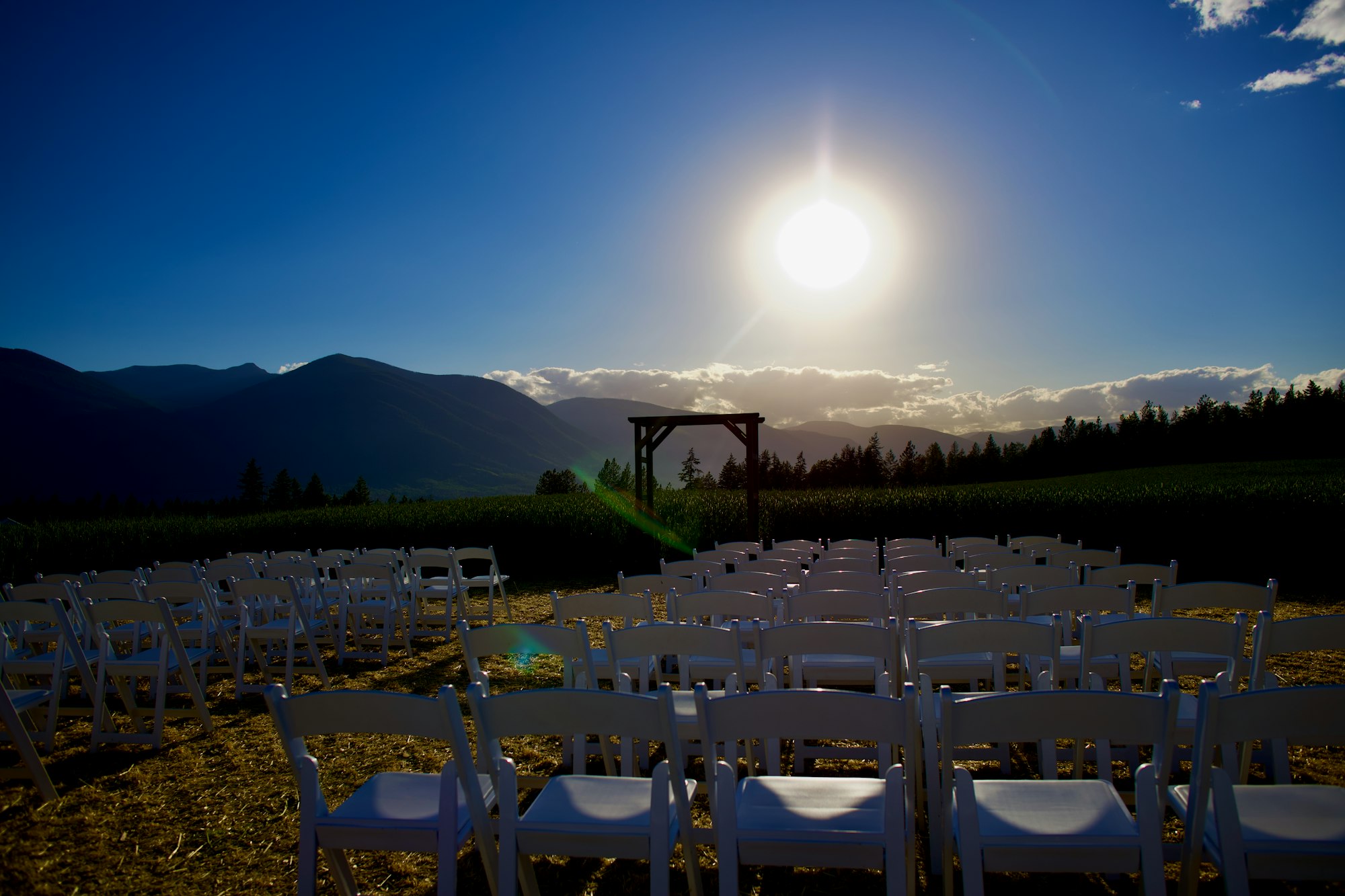 Rows of empty white folding chairs facing a timber arch on a hillside, with the sun sitting low over the ranges behind it